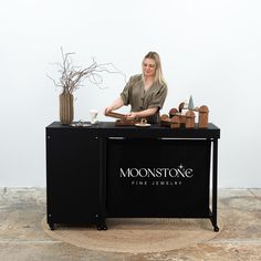 a woman standing behind a black counter with items on it and a vase in the background