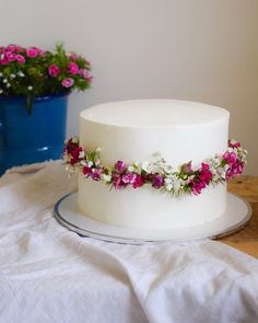 a white cake with pink and white flowers on it sitting next to a blue potted plant