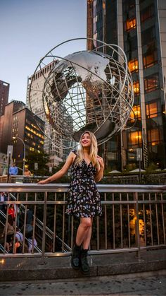 a woman standing in front of a large metal globe with people walking around it and buildings behind her