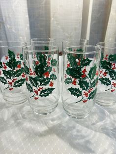 four glasses with holly and red berries on them sitting on a white table cloth next to a window