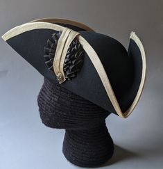 "This military style three cornered tricorn hat is made from top quality wool felt. The edges have been trimmed even and the sides held up or \"cocked\". It has been trimmed around the brim with gold fold-over braid. It is also available with black or white braid. Plain untrimmed hats are listed separately. Gold tone of edging may vary due to availability. Attached is a 3.5 \" black and gold, pleated double cockade with gold fleur de lis button below. Other buttons are available in the variation Formal Black Brimmed Costume Hats And Headpieces, Formal Black Costume Hats With Curved Brim, Black Formal Costume Hat With Curved Brim, Formal Black Brimmed Costume Hat, Black High Crown Hat For Formal Occasions, Black High Crown Costume Hat For Formal Occasions, Formal Black High Crown Costume Hat, Black Fitted High Crown Costume Hat, Fitted Black Hat With Structured Crown