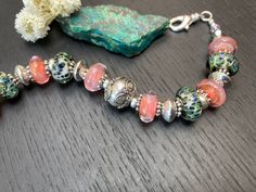 "Perfect for spring and summer, this adjustable bracelet features gorgeous artisan lampwork glass beads that are peach pink on one side and speckled green and blue on the other, combined with pink glass rondelles, green and brown speckled glass rondelles, and accented with gleaming with sterling donuts and Bali granulated sun and daisy spacers, and green and pink Swarovski crystals. Finished with a Silver plated lobster clasp an extension chain with charm. Fits as an approximately 6 7/8 inch bra Handmade Peach Bracelets With Round Beads, Handmade Peach Beaded Bracelets For Gift, Handmade Peach Beaded Bracelets As Gift, Adjustable Peach Beaded Bracelet As Gift, Adjustable Coral Bracelets As A Gift, Adjustable Peach Beaded Bracelets As Gift, Handmade Peach Bracelet Gift, Peach Round Beads Bracelet As Gift, Adjustable Pink Bracelets