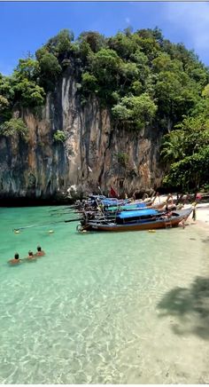 people are swimming in the clear blue water near boats on the beach and cliffs behind them