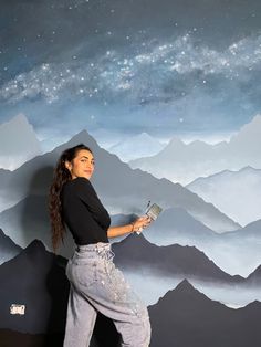 a woman standing in front of a wall painting with mountains and stars painted on it