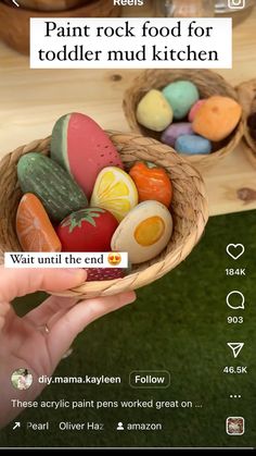 a person holding a basket full of fake food in front of a screen with the words paint rock food for toddlers mud kitchen