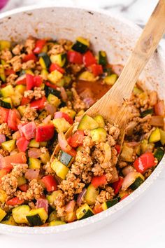 a white bowl filled with meat and vegetables next to a wooden spoon on top of a table