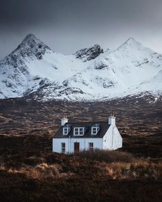 a small white house in the middle of a field with snow covered mountains behind it