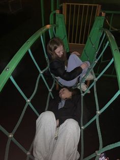 two people are sitting in a green swing set at the park, looking down on them