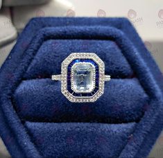 an engagement ring in a blue velvet case with diamonds on it and the center stone is surrounded by smaller round brilliant cut diamonds