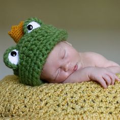 a baby wearing a green crocheted hat with googly eyes laying on top of a blanket