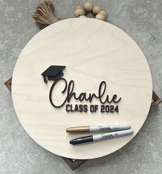 a round wooden sign that says charlie class of 2014 with two pens on it and some beads