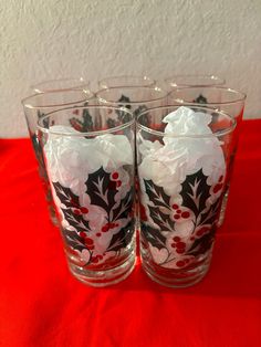 four glass cups with holly and white flowers on them sitting on a red tablecloth