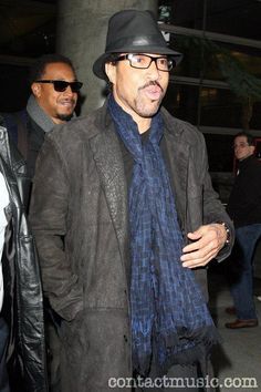 a man in a hat and scarf walking down the street with another man behind him