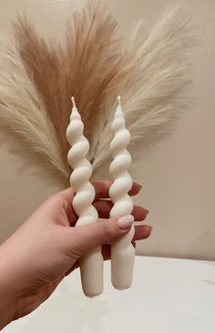 a hand holding two white candles in front of a fake plant with long brown feathers