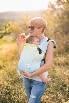 a woman holding a baby in her sling while walking through the grass with an apple