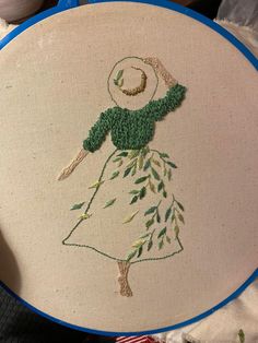 a hand embroidered picture of a woman in a green dress and hat on a blue hoop