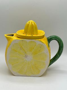 a yellow and green teapot with a lemon slice on it's side, sitting on a white surface