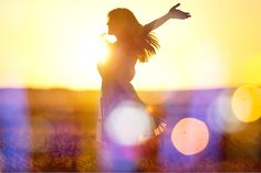 a woman standing in a field with her arms out and the sun setting behind her