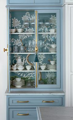 a blue china cabinet with gold handles and glass doors, decorated with white floral wallpaper