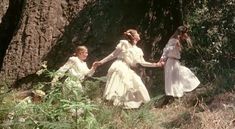 two women in white dresses are holding hands with another woman standing next to them near a tree