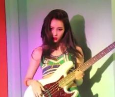 a woman holding a bass guitar in front of a purple and green wall with light coming from behind her