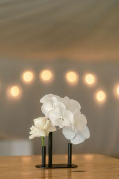 two black vases with white flowers are on a wooden table in front of lights