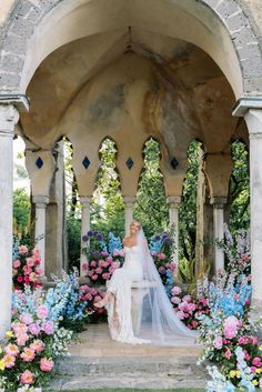 a woman in a wedding dress is standing under an arch with flowers on the ground