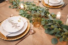 a wooden table topped with white plates and gold rimmed dishes covered in greenery