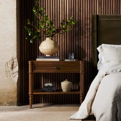 a bed with white sheets and pillows next to a wooden shelf holding a potted plant