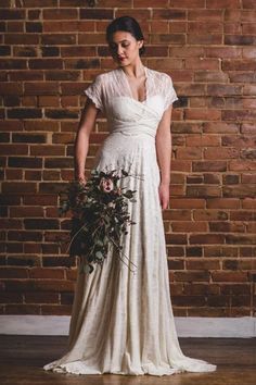 a woman standing in front of a brick wall wearing a white dress and holding a bouquet