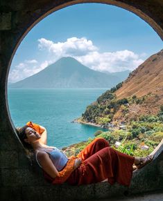 a woman laying down in a stone window sill looking out at the ocean and mountains