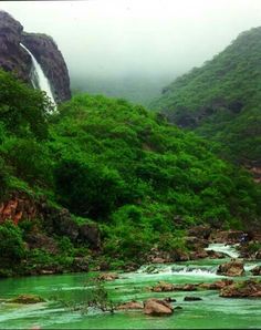 a river running through a lush green forest next to a tall rock formation with a waterfall in the background