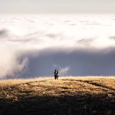 two people standing on top of a hill with clouds in the background and one person holding an umbrella