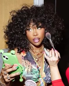 a woman with curly hair holding a cell phone and looking at her makeup brush in front of her face