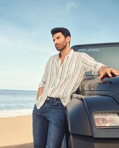 a man leaning on the hood of a black car near the beach with his hands in his pockets