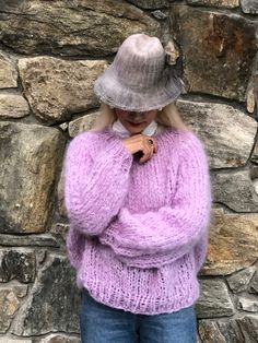 a woman standing in front of a stone wall wearing a purple sweater and gray hat