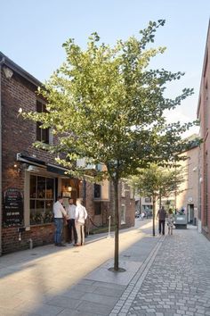 people are walking down the sidewalk in front of a tree and brick building with shops on it