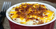 a red bowl filled with mashed potatoes covered in bacon next to a fork and knife