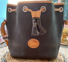 "Here is a stunning vintage Dooney and Bourke handbag in a two tone chocolate and peanut butter color! This beautiful bag is made of all weather leather and has the duck emblem on the front! It has a drawstring closure with tassel detail! There is an attached key holder on the inside to attach your keys to. The hardware is all solid brass. This purse also features an adjustable strap! Made in USA Approximately 13.75\" long 10\" deep 11\" from the top of the strap to the purse 6\" wide Very good Vintage Dooney And Bourke, Metal Napkin Rings, Chocolate And Peanut Butter, Chocolate Leather, The Duck, Hand Mirror, Dooney And Bourke, Dooney & Bourke, Key Holder