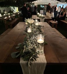 a long table with candles and greenery on it in front of people at an outdoor event