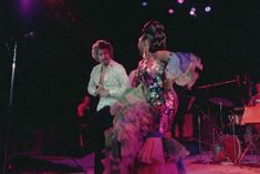 two people are dancing on stage in front of microphones and music instruments, one is wearing a colorful dress