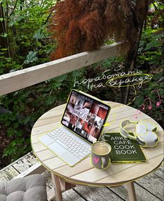 a laptop computer sitting on top of a wooden table next to a book and cup