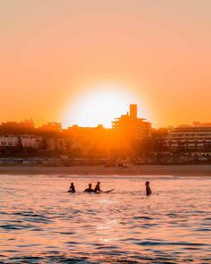 three people in the water at sunset with buildings in the backgrouds and one person on a surfboard