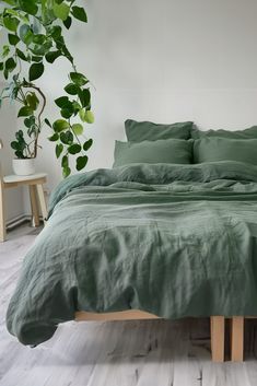 a bed with green sheets and pillows next to a potted plant in a white room