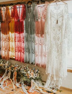 wedding gowns and dresses hanging in front of a window with flowers on the floor