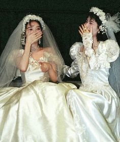 two women dressed in wedding gowns sitting next to each other and covering their mouths