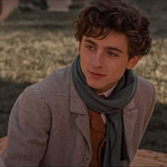 a young man wearing a scarf and jacket sitting at a table in front of some grass