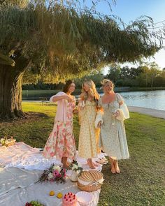 three women in dresses standing around a table with food on it near water and trees