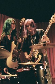 three women with green hair are playing guitars in front of red curtained walls and drums