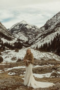 a woman in a wedding dress and fur stole standing on the side of a mountain